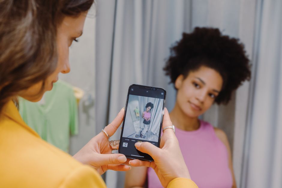 Two women enjoy a shopping day, capturing a moment in the fitting room with a smartphone.