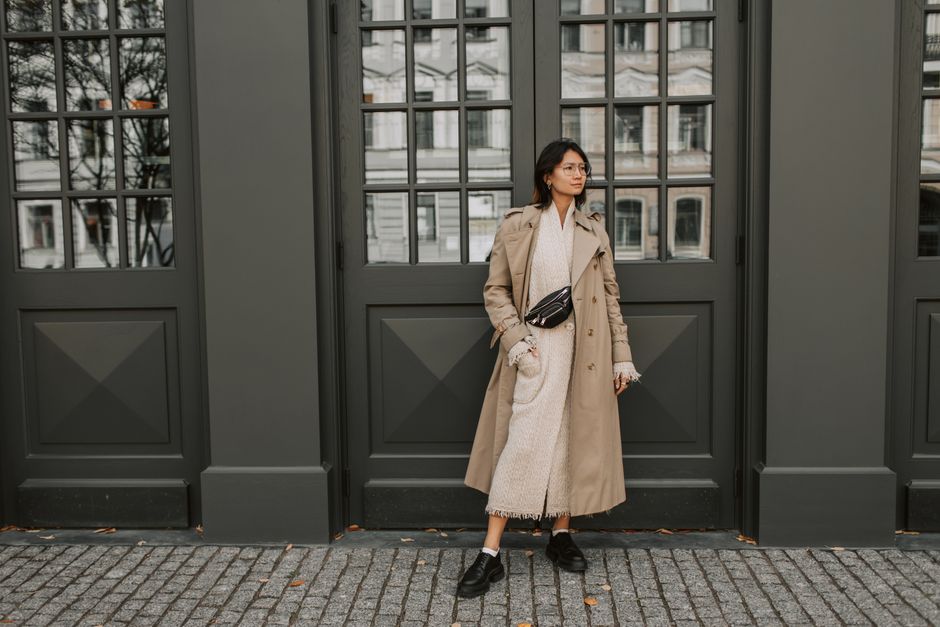 A fashionable woman poses confidently in a trench coat against an urban backdrop, showcasi
