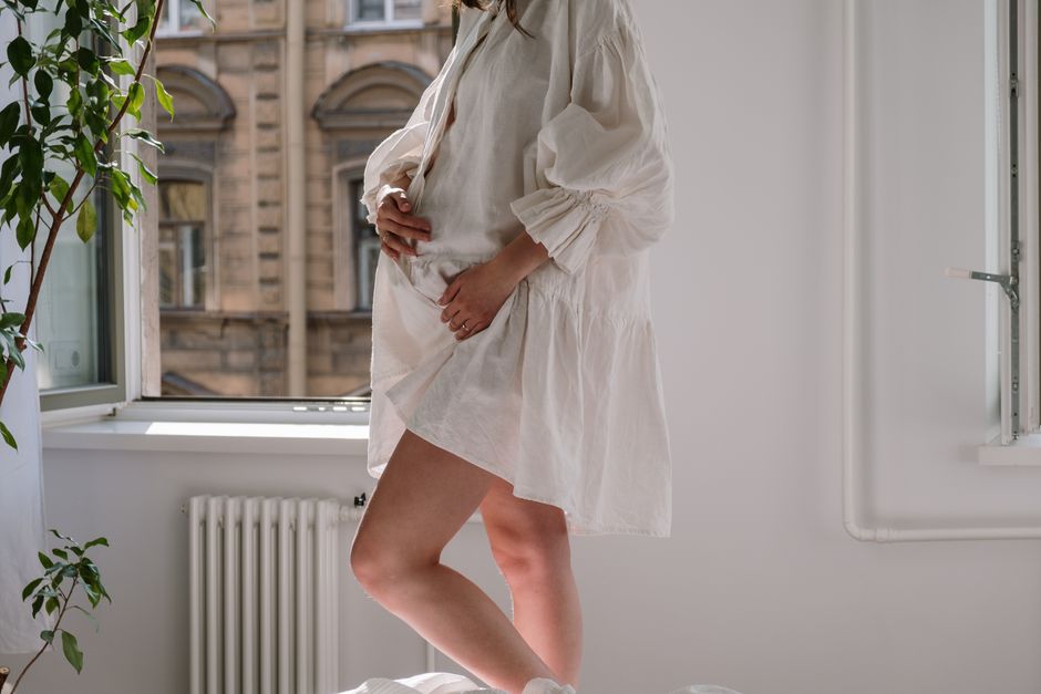 A pregnant woman stands by a window in a white dress, gently holding her belly.