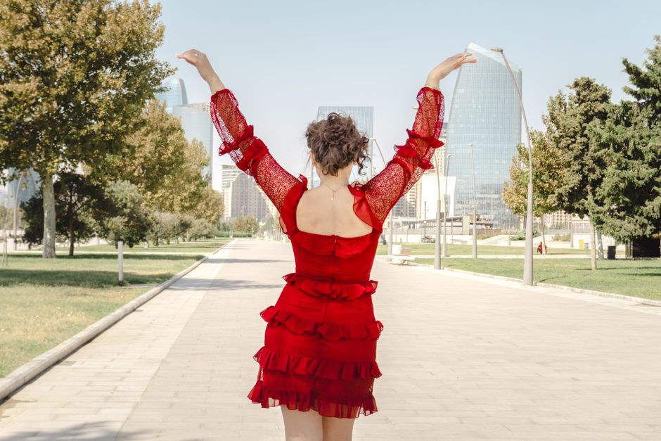 A woman in a red dress joyfully raises her arms while standing in an urban park with moder