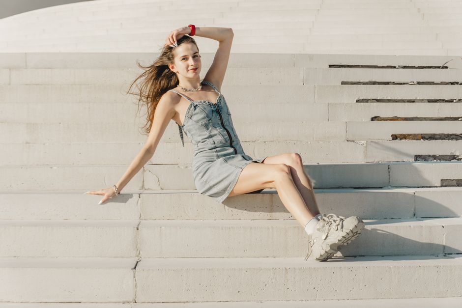 A young woman in a denim dress relaxes on outdoor steps, enjoying a sunny day.