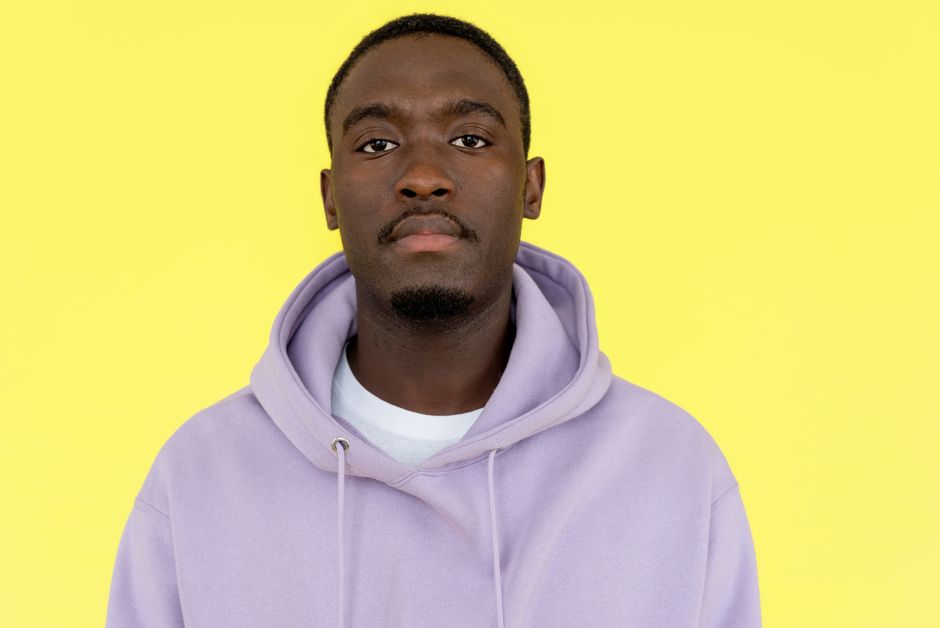 Close-up portrait of a man wearing a purple hoodie against a vibrant yellow background with neutral expression.