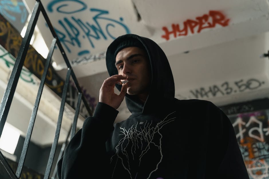 Man wearing a black hoodie smoking in a graffiti-covered staircase, urban and edgy atmosphere.