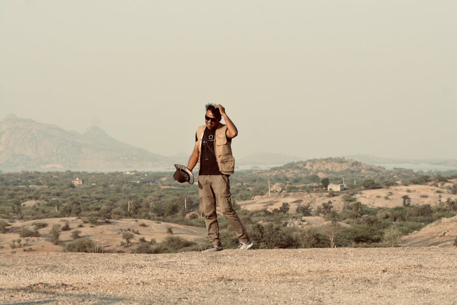 A man in adventure attire stands in a desert with a mountainous background.