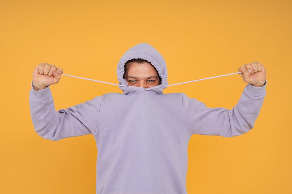 Man playfully pulling hoodie strings over face on vivid background.
