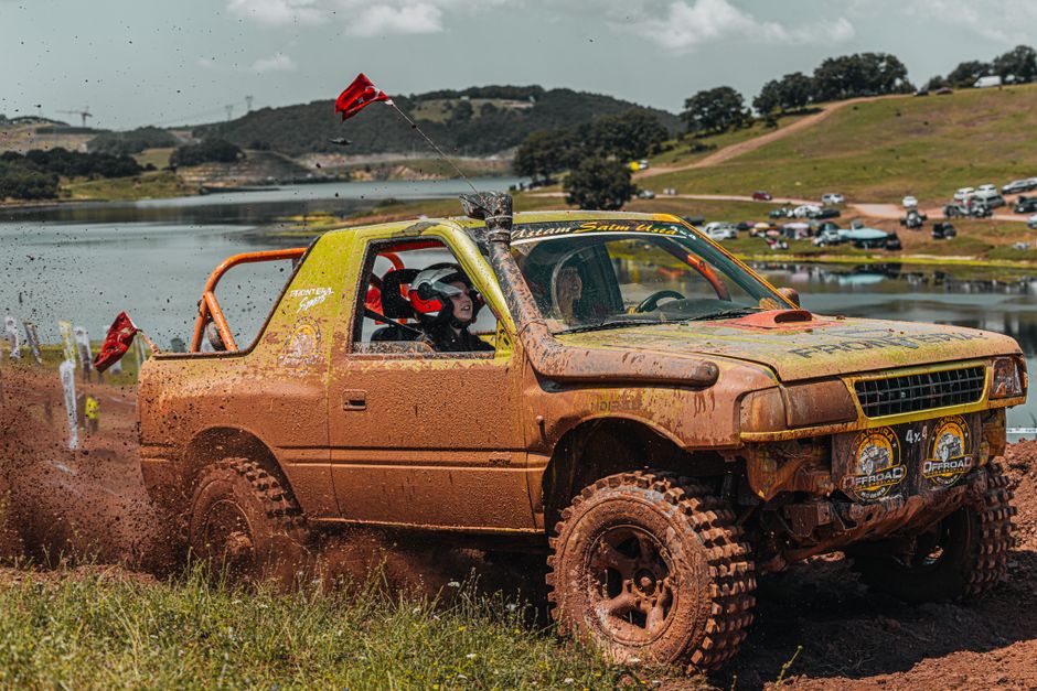 Off-road truck racing through muddy terrain with driver in safety gear during daytime.