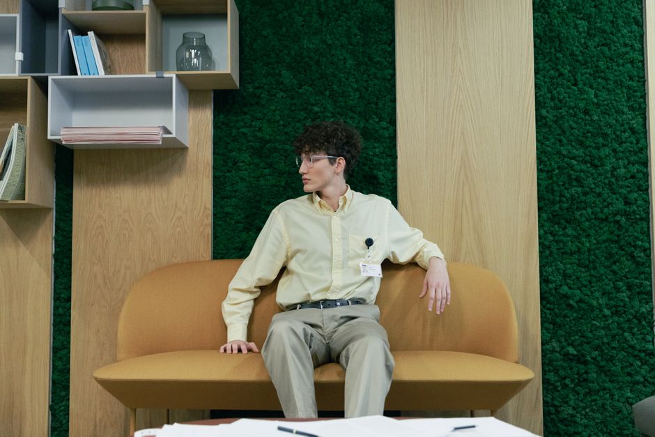 Young adult with curly hair and eyeglasses seated on a modern sofa in an indoor office setting.