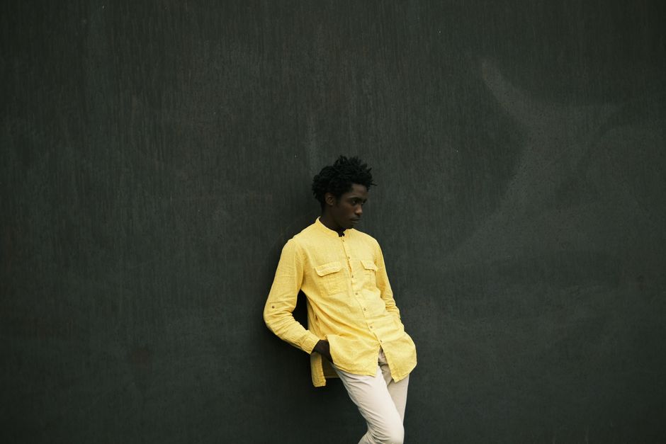 Stylish young man wearing a yellow shirt poses against a dark wall.