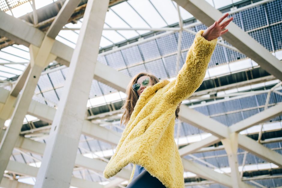 Fashionable woman in yellow faux fur coat and sunglasses poses under modern architecture.