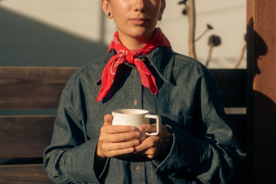 Fashionable young person with coffee in sunlight wearing denim and bandana.