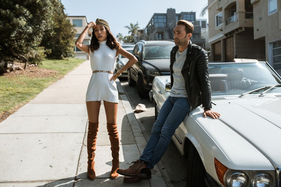 Fashionable couple in urban setting, posing by cars on a sunny street.