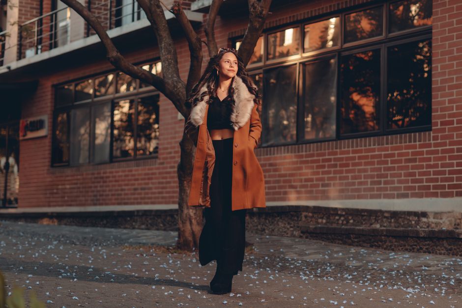 Fashionable woman in coat walking on urban sidewalk during sunset.