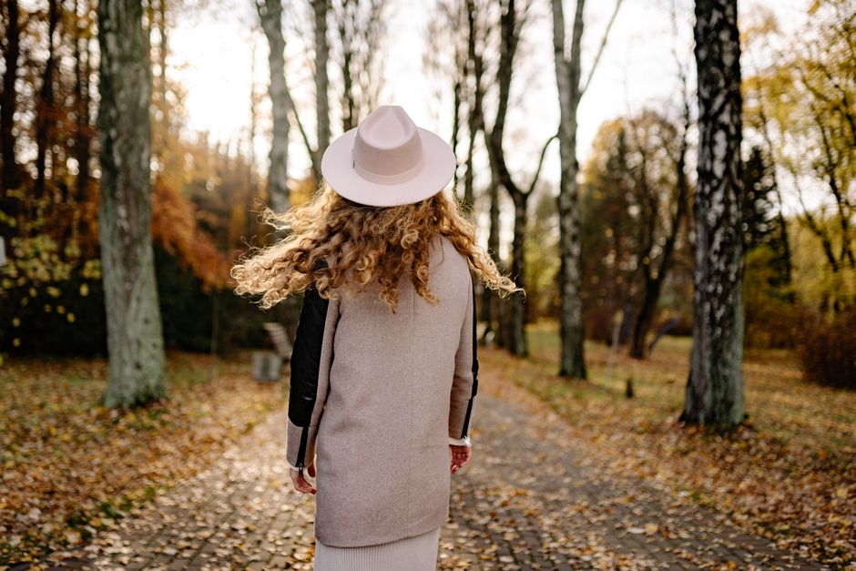 A woman with curly hair in a hat walks down a leaf-covered park path in autumn.