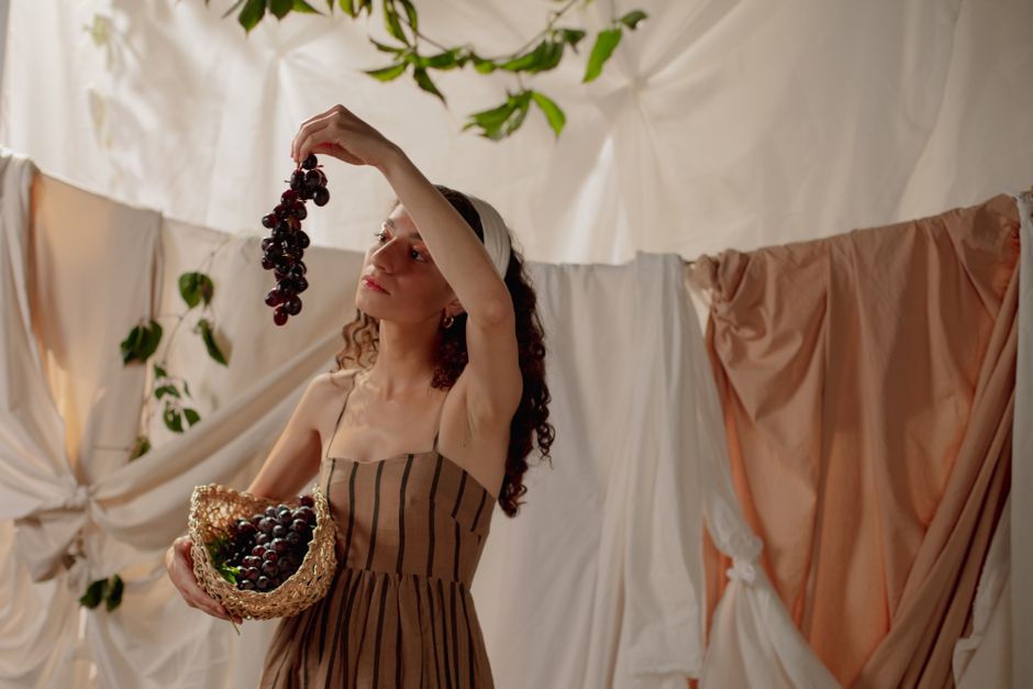A woman in a striped dress holds grapes in a rustic, draped setting.