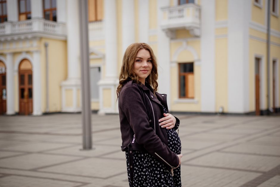 A pregnant woman in a black jacket stands smiling in an urban environment.