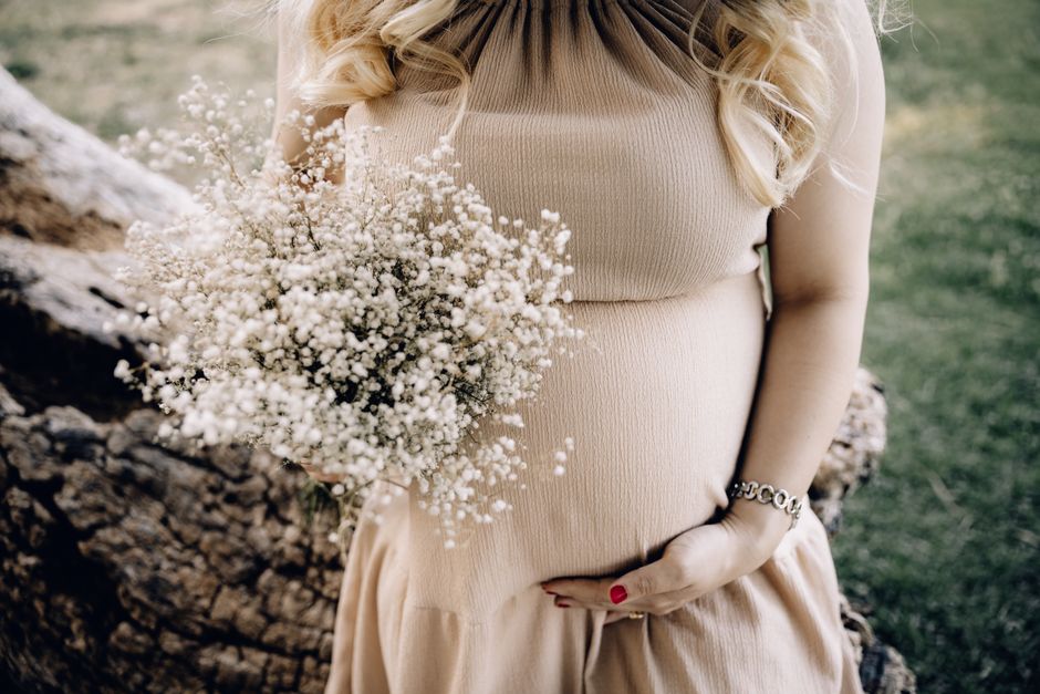 Pregnant woman in beige dress holding baby's breath bouquet outdoors.
