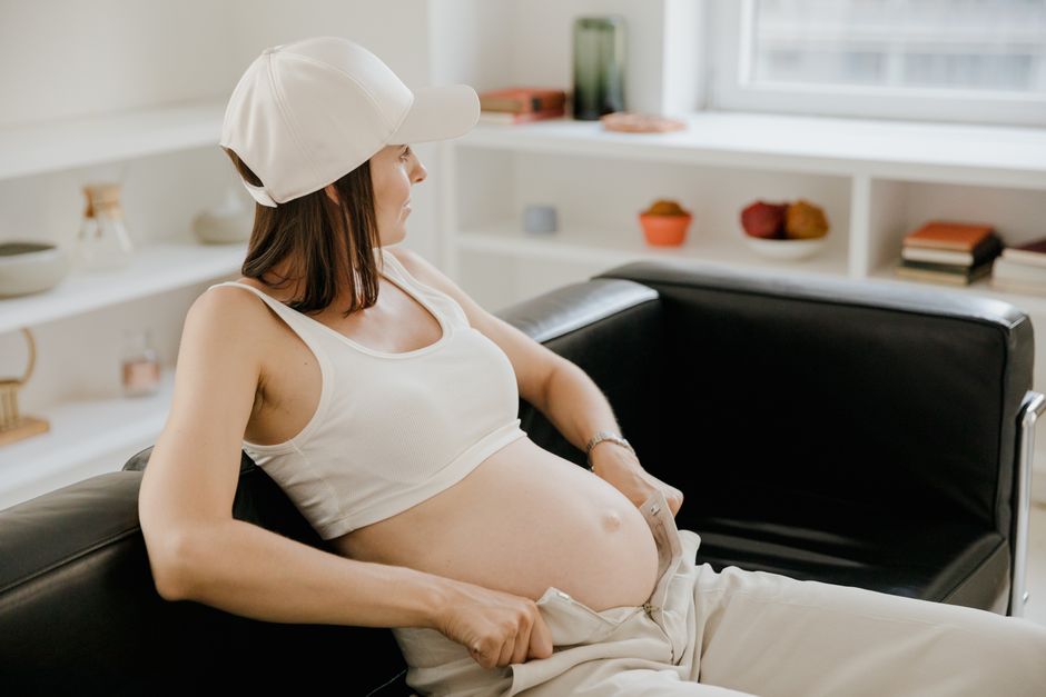 Pregnant woman in a white cap rests on a black sofa, showcasing her baby bump in a bright indoor setting.