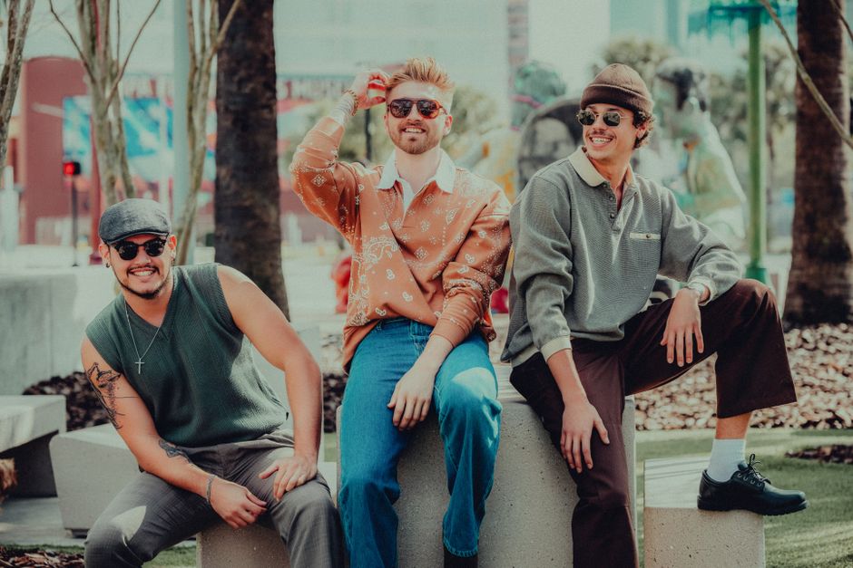 Three stylish men sitting outdoors in urban area, wearing sunglasses and smiling.