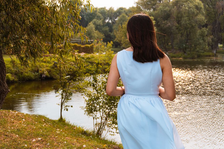 A woman in a blue dress stands elegantly by a serene lake in a park setting.