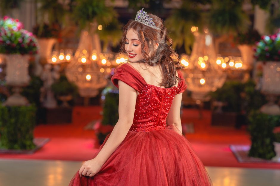 Woman wearing a sparkling tiara and red gown twirling at an elegant indoor celebration.