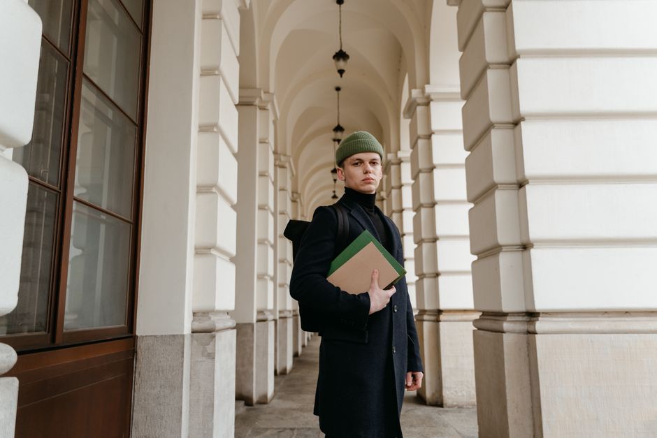 A fashionable man in a black coat and green beanie holds books in an elegant archway.