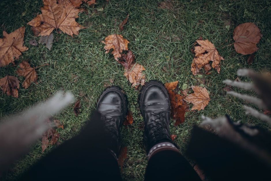 Top view of black boots on grass covered with autumn leaves, showcasing fall vibes.