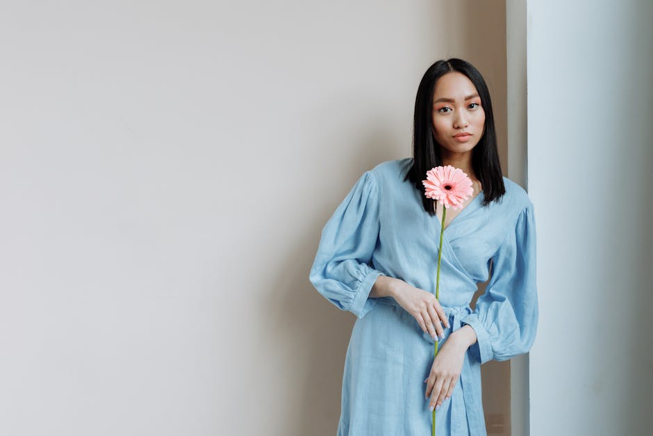 Elegant young Asian woman in a blue dress holding a pink gerbera, posing with confidence.