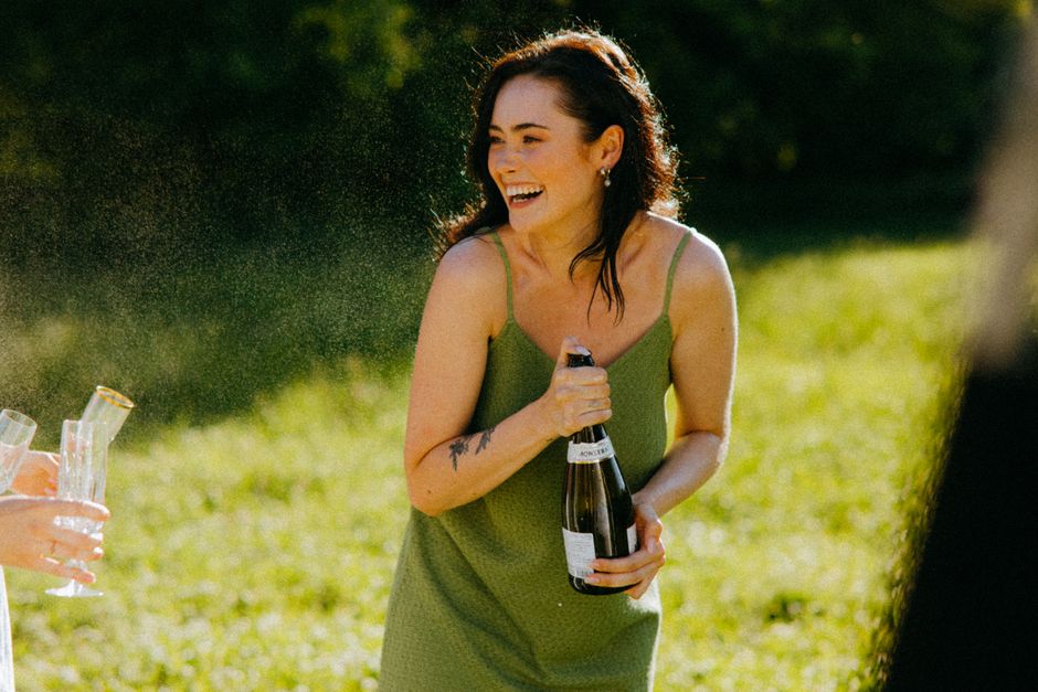 Woman in green dress joyfully opens champagne outdoors on a sunny day.