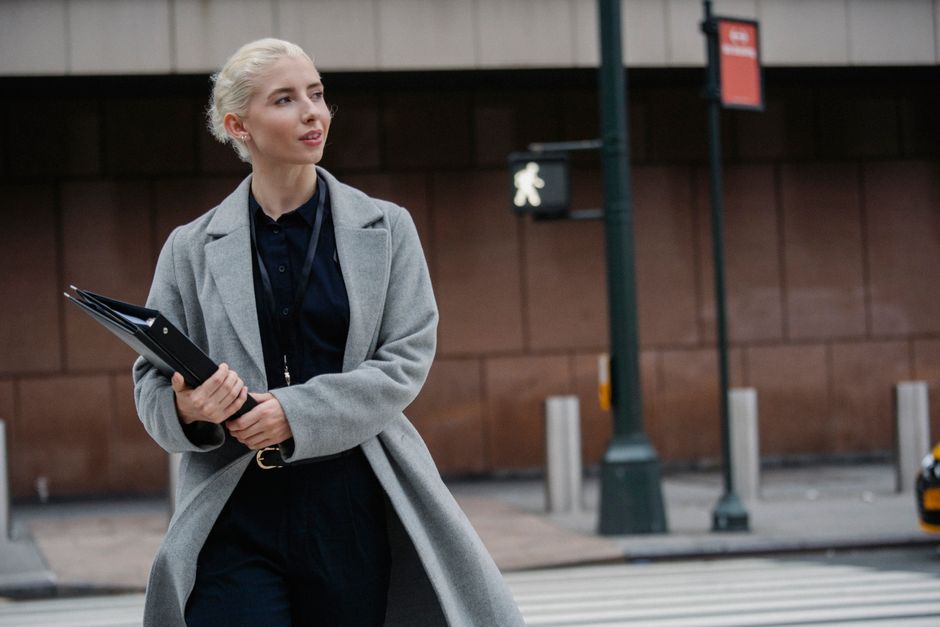 Content young businesswoman wearing trendy gray coat carrying black folders and crossing zebra on busy street while looking away