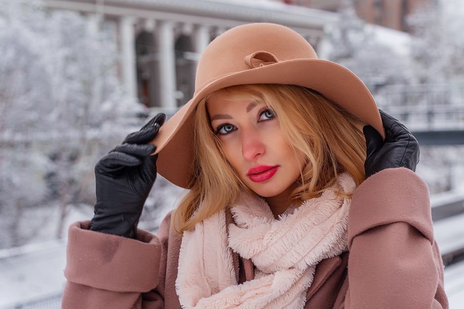 Woman in a fashionable winter coat and hat poses outdoors in a snow-covered setting.
